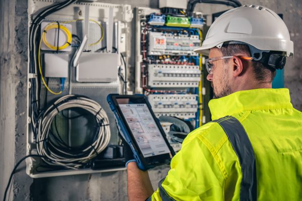 Man, an electrical technician working in a switchboard with fuses, uses a tablet. Man, an electrical technician working in a switchboard with fuses. Installation and connection of electrical equipment. Professional uses a tablet.